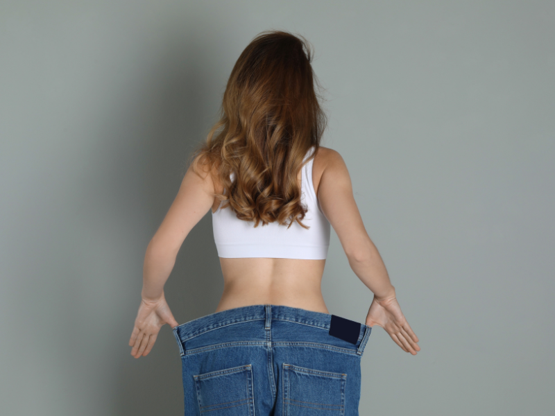 Frau ab 40 steht mit dem Rücken da und zieht die zu große Jeans etwas weg, damit man erkennt wieviel sie abgenommen hat und Bauchfett verloren hat und jetzt ihr Wohlfühlgewicht hat